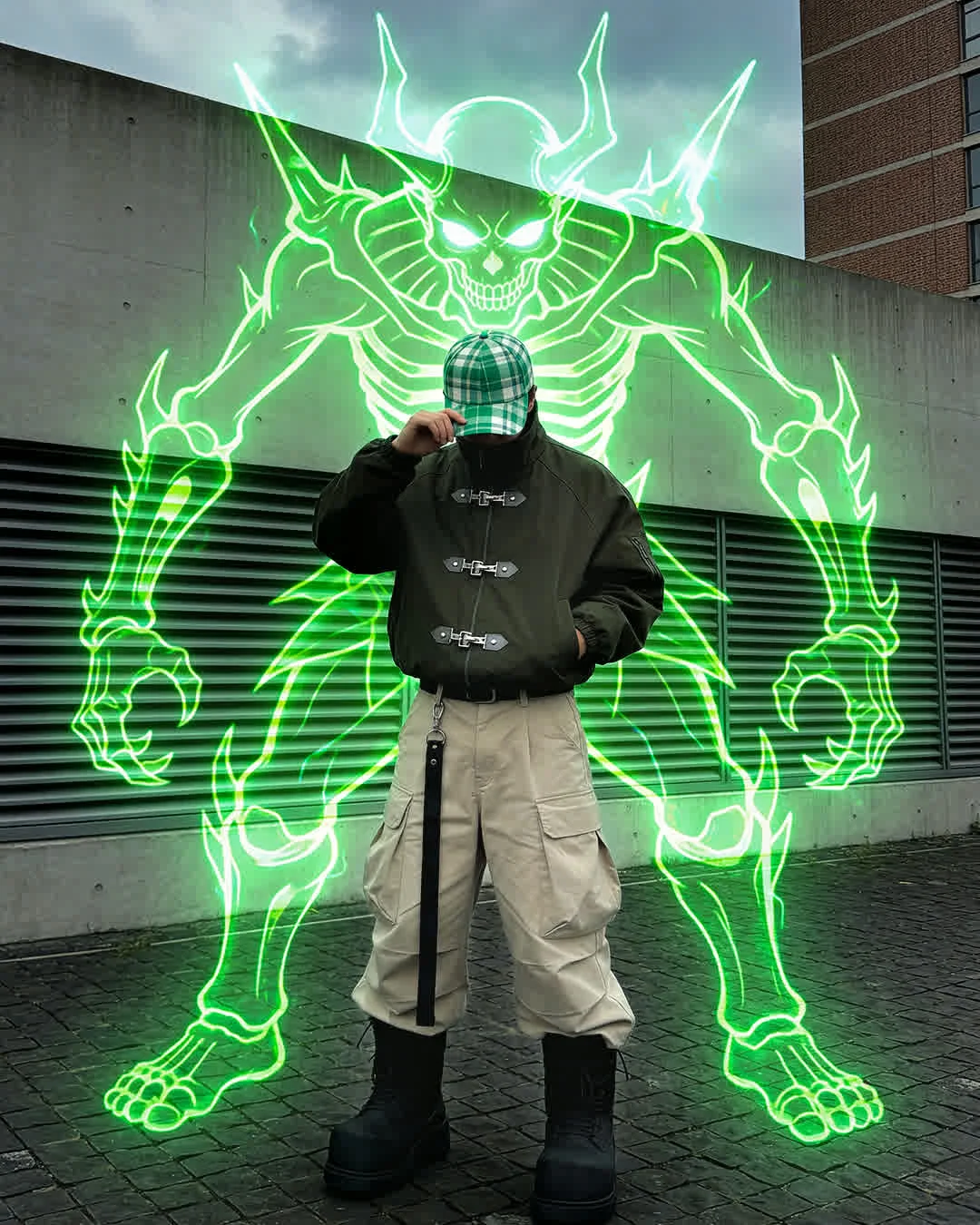 Create a dynamic digital composite that blends gritty urban streetwear photography with cyberpunk anime visuals. The central figure is a young man dressed in tactical modern fashion, wearing an olive green jacket featuring distinctive silver metal clasp closures, loose beige cargo trousers, and chunky black combat boots. He wears a green and white plaid baseball cap pulled low to obscure his face, adjusting the brim with one hand while the other rests in his pocket. Looming behind and around him is a towering, neon-green skeletal avatar. This spectral entity resembles a demonic "Stand" or summon, composed of glowing electric green outlines that form a horned skull, ribcage, and spiky skeletal limbs. The setting is a gloomy, overcast urban plaza with grey concrete walls and cobblestone pavement. The composition should contrast the realistic, high-fidelity textures of the clothing against the vibrant, luminous vector-art style of the neon skeleton.