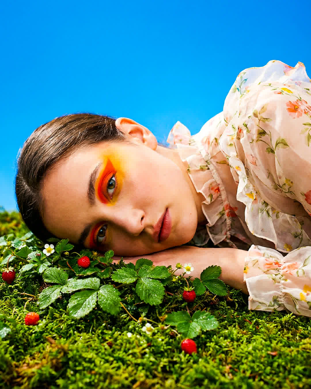 Create a close-up editorial shot of a woman lying on a vibrant green surface, her head resting gently on the ground. She is wearing a delicate, sheer floral blouse with soft ruffles, and her makeup is bold with orange and yellow hues around her eyes, adding a striking contrast to her natural beauty. The background is a bright blue sky. The lighting should be soft and bright, highlighting the sharp details of the model's facial features and the delicate textures of the blouse and floating elements. The composition should emphasize the dreamy, serene atmosphere, with a balance between the rich greens of the surface and the freshness of the strawberries and flowers.
The image should have high contrast and vibrant colors, evoking a playful, surreal mood.