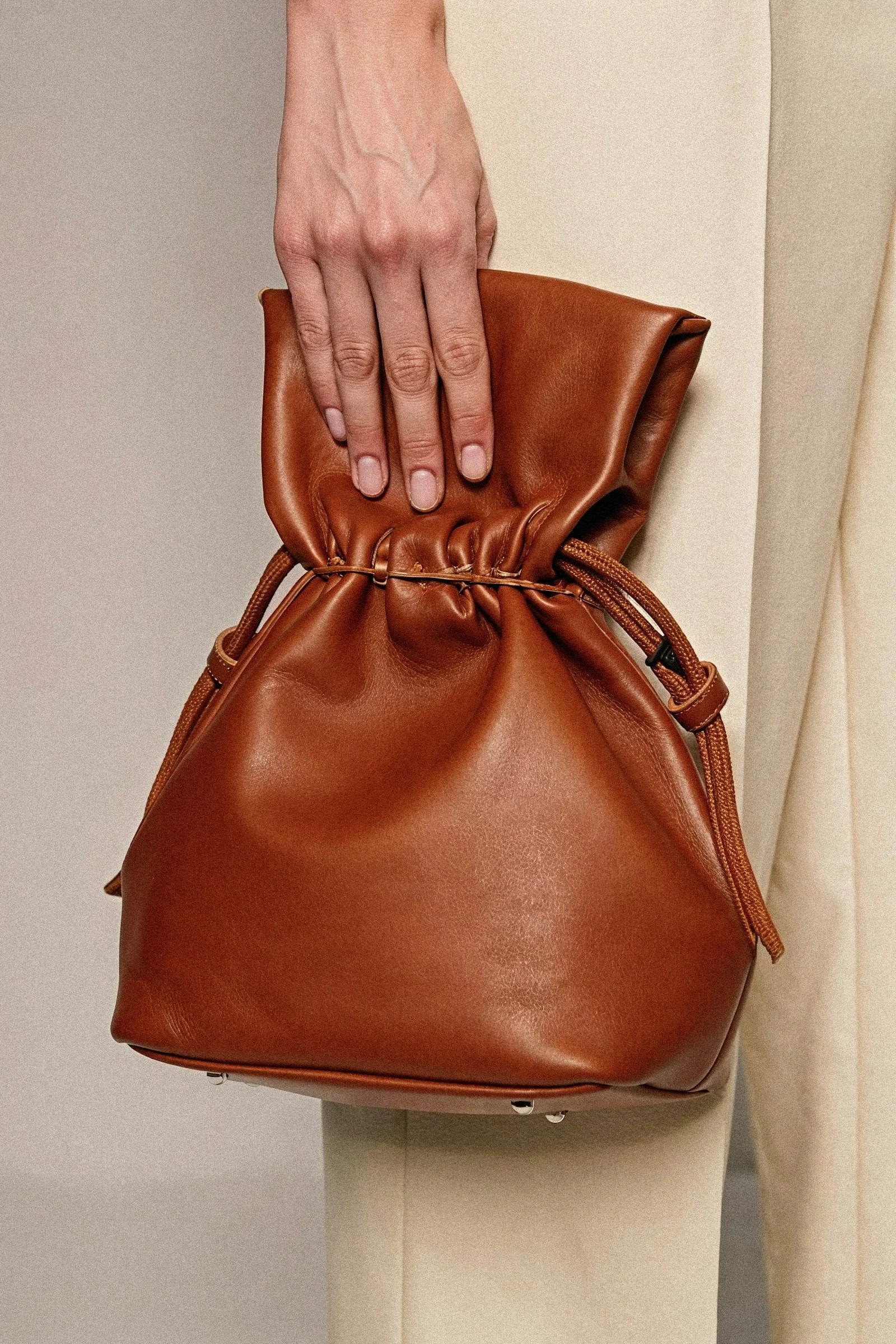 Close-up of a hand holding a caramel brown leather drawstring bag with a slouchy design, perfect for casual yet chic outfits.
