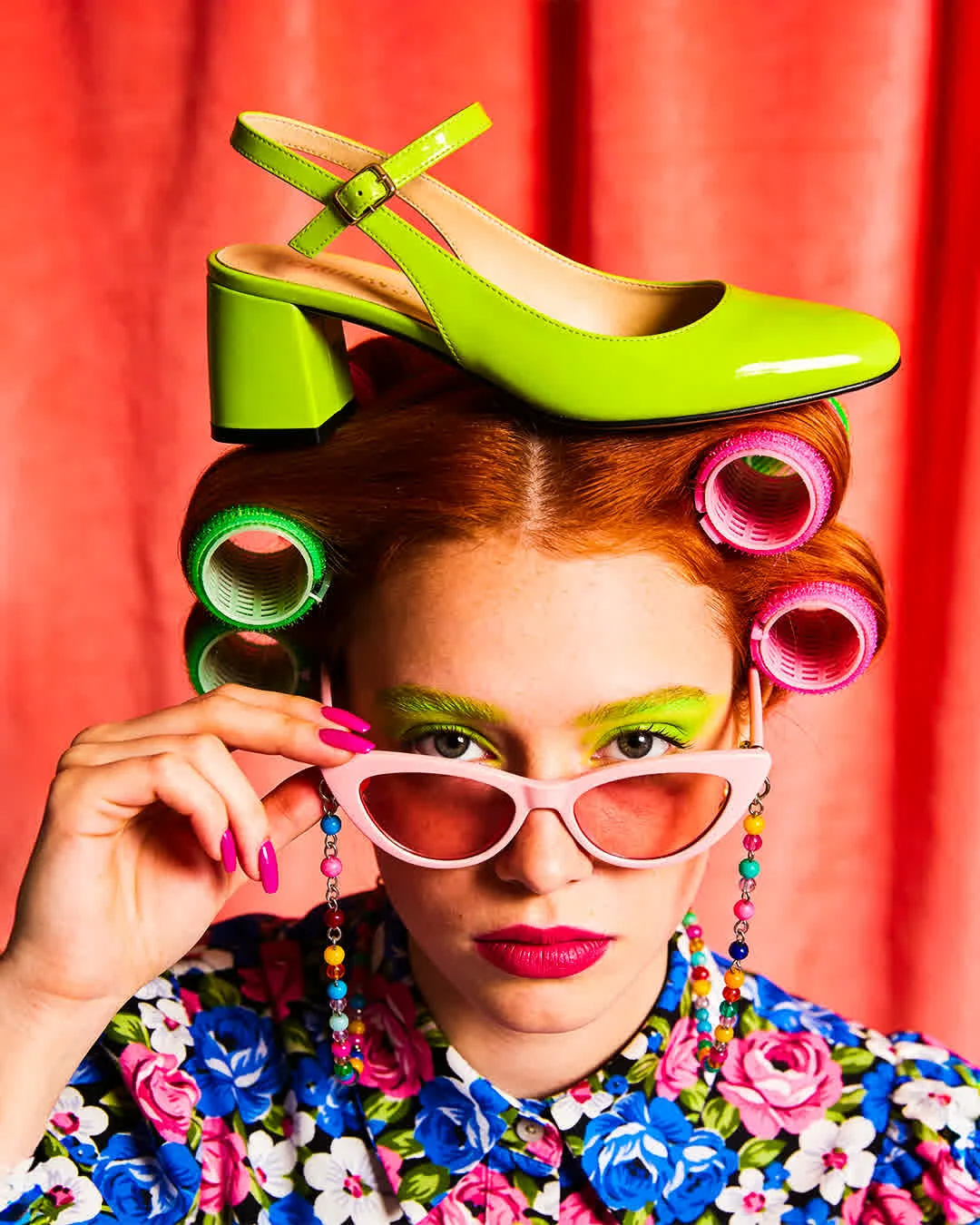 Create a vibrant, surreal pop-art fashion portrait that blends retro 1950s domestic aesthetics with high-saturation maximalism. The central figure is a young woman with copper-red hair styled in vintage victory rolls, accented by large, colorful velcro hairrollers in neon pink and green. Her makeup is bold and theatrical, featuring a wash of electric lime-green eyeshadow and vivid fuchsia lipstick. She is peering over the rim of pale pink cat-eye sunglasses with rose-tinted lenses, holding the frame with one hand displaying a bright pink manicure. The glasses feature playful, múlti-colored beaded chains dangling down. She wears a busy, vintage-style blouse with a dense floral print of blue, pink, and white roses The most stnking surreal element is a glossy, neon lime green patent leather block-heeled shoe balanced playfully on top of her head like a hat. The background is a textured, coral-red draped curtain.