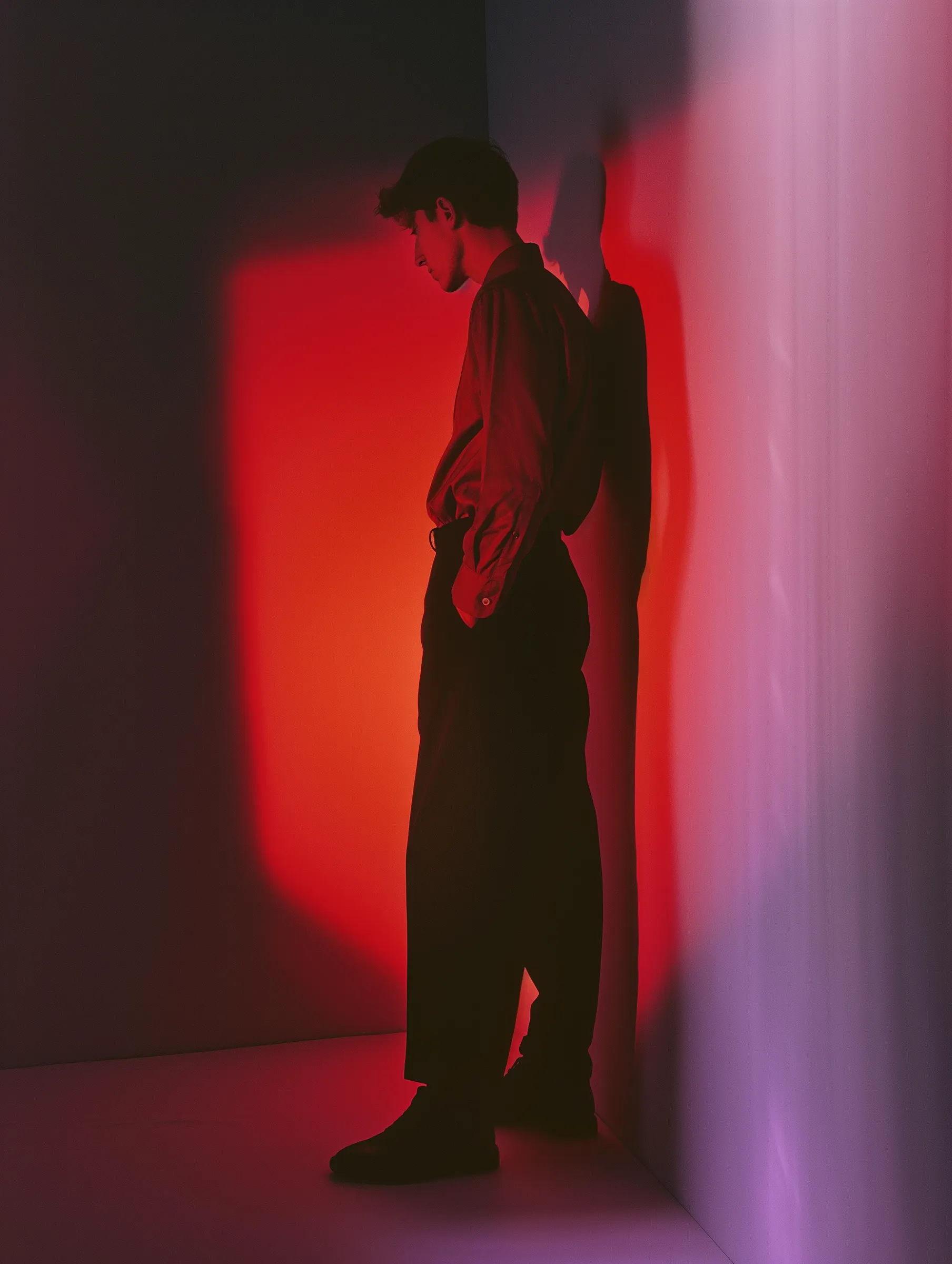 A person in a dimly lit room with dramatic red lighting, leaning against a wall, creating a striking contrast with shadows.
